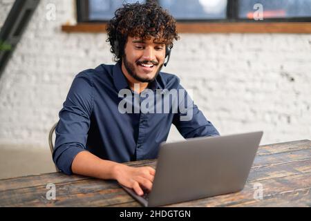 Smiling ethnic Indian man wearing wireless headset using laptop for virtual connection, talking with colleagues, Indian male student involved webinar. Latin employee of customer service support Stock Photo