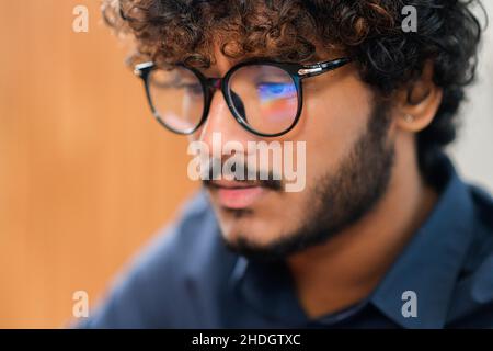 Selective focus at the reflection of screen on the glasses of Indian male employee, concentrated multi ethnic male freelancer in glasses using laptop, indian latin curly developer guy coding Stock Photo