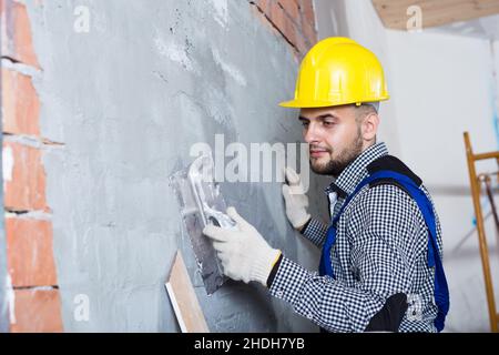 Smiling male in the helmet is plastering the wall Stock Photo