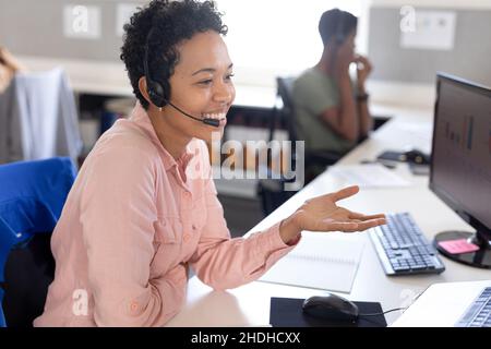 Smiling biracial female customer representative gesturing while talking through headset in office Stock Photo