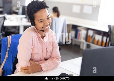 Biracial female customer representative smiling while talking through headset in office Stock Photo
