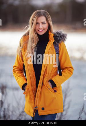 Casual portrait of young woman in warm orange yellow winter jacked, zipper down, smiling, blurred snow covered landscape behind her Stock Photo