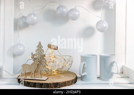 A wooden figurine of deer and spruce against the background of a metal decorative basket, two mugs of tea, a silver garland of threads on the wall dec Stock Photo