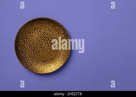 Textured round golden plate on blue violet linen paper background. Color of the year 2022 concept. Modern crockery for trendy food design. Copy space. Stock Photo