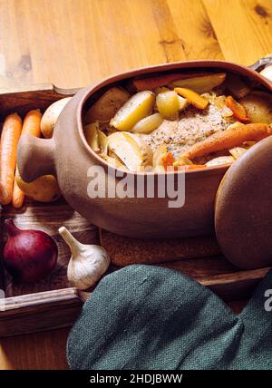 Side view of terracotta clay cooking pot with slow cooked pork roast and vegetables inside on wood tray and wooden table, surrounded by raw organic. Stock Photo