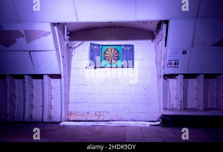 An old darts board, in a tunnel underground, that has been well used and is lighted on the wall. Stock Photo