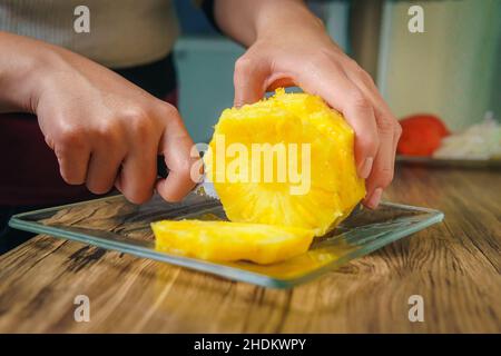 Close-up of a young woman's hands cutting a juicy pineapple with a chef's knife on a wooden table in a kitchen. Stock Photo