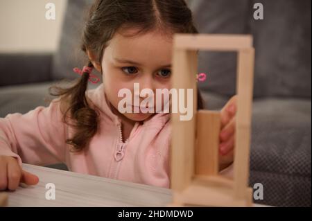 Close-up portrait of adorable Caucasian child, preschool girl in pink sweatshirt concentrated on building wooden structure with wooden blocks. Board g Stock Photo