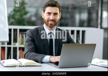 Portrait handsome successful caucasian bearded businessman, ceo of company, top manager, wearing formal suit, sitting at table with laptop in modern office, looking at the camera and smiling friendly Stock Photo