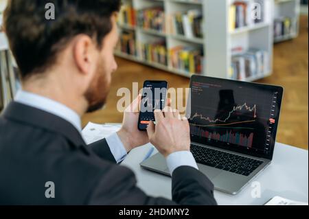 Exchange market. Smart man investor and crypto trader, using laptop and cellphone, analyzes charts of trading in stock market and digital cryptocurrency exchange,conducts analysis,trading crypto coins Stock Photo