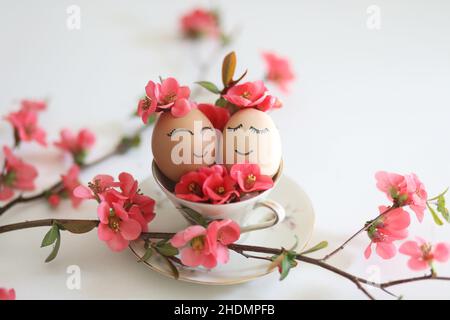 Creative easter eggs with cute face, sleepy eyes and spring flowers in tea cup on white background. Happy Easter concept, mockup, copy space.  Stock Photo