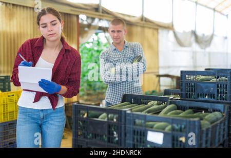 Female farmer working with papers in warehouse, male worker carrying crates with cucumber Stock Photo