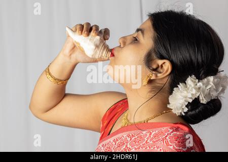 A beautiful Indian woman in red saree blowing conch shell on white background Stock Photo