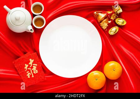White plate on red satin cloth background with tea set, ingots, red bag (word means wealth), oranges and red envelope packets or ang bao(word means au Stock Photo