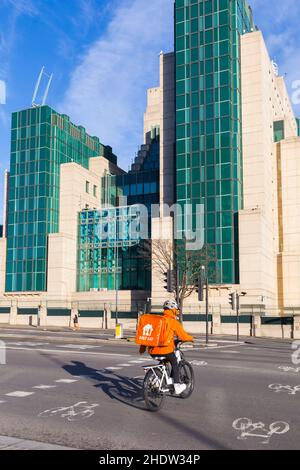 A Just Eat delivery rider on a bicycle crosses on a cycle lane at a junction, with MI6 building on the other side at Vauxhall, London UK in December Stock Photo