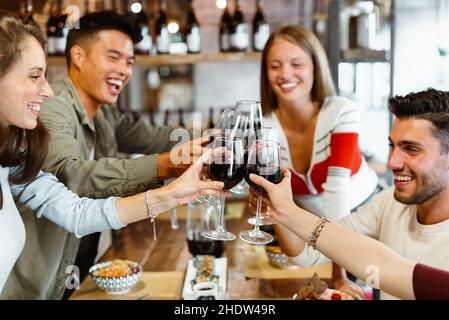 Diverse group of multicultural friends celebrating in restaurant together laughing and joking as they toast each other with glasses of red wine Stock Photo