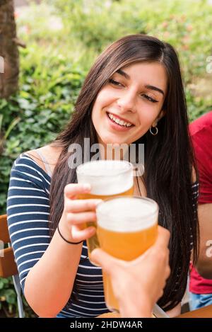 Cheerful female clinking glass of refreshing beer with unrecognizable friends while celebrating reunion during picnic in park Stock Photo