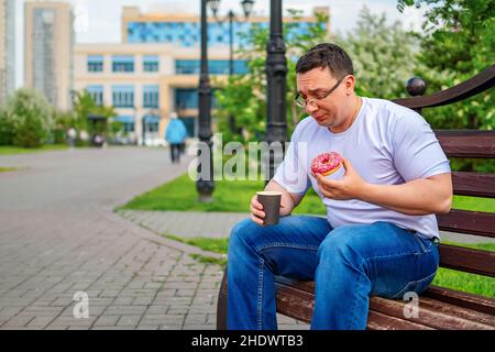 a despondency, puzzled, sad man with glasses is sitting in a city park, having lunch, eating a sweet donut, a bun and holding a cup of coffee, tea Stock Photo