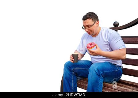 a sad, puzzled, sad man with glasses is sitting on a bench, having lunch, eating a sweet donut, a bun and holding a cup of coffee, tea Stock Photo