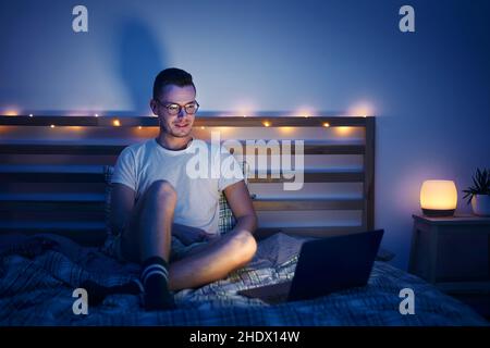 Lonely man with eyeglasses sitting on bed in bedroom and using laptop at night. Face illuminated from computer display. Stock Photo