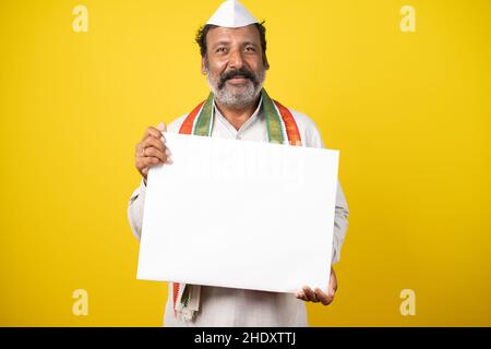 Portrait of happy smiling Indian Poliician holding Empty white sign board by looking at camera on yellow studio background. Stock Photo