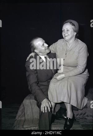 1956, historical, happy elderly couple sit together on a rug covered box for their photo. Both well-dressed, the man is in a suit & tie and the lady in a dress, smiling, sitting on the man's lap. Organisers of a May Day carnival, inside a church hall, England, UK. Stock Photo