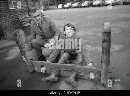 1986, historical, outside a school building, a male teacher and teenage boy demonstrate an ancient wooden 'foot and hand lock', known as the stocks, an instrument of corporal punishment, used to restrain and humiliate offenders. A key part of the stocks was the element of public punishment. It's last recored use in Britain was in 1872. The stocks differed from the pillory, which locked the head of the offender forcing them to stand and where custom dictated that their head and beard were shaved. Stock Photo