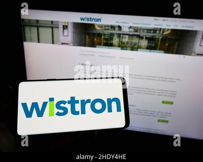 Person holding mobile phone with logo of Taiwanese electronic company Wistron Corporation on screen in front of web page. Focus on phone display. Stock Photo