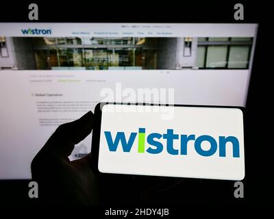 Person holding cellphone with logo of Taiwanese electronic company Wistron Corporation on screen in front of webpage. Focus on phone display. Stock Photo