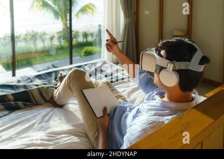 Young Asian man lying on bed and using virtual reality glasses for business meeting during his travel summer vacation holiday. Stock Photo