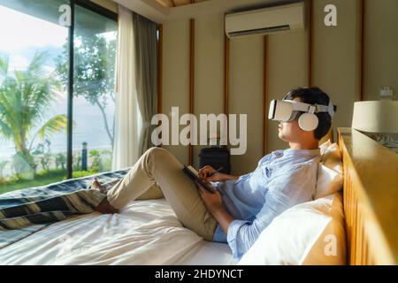 Young Asian man lying on bed and using virtual reality glasses for business meeting during his travel summer vacation holiday. Stock Photo