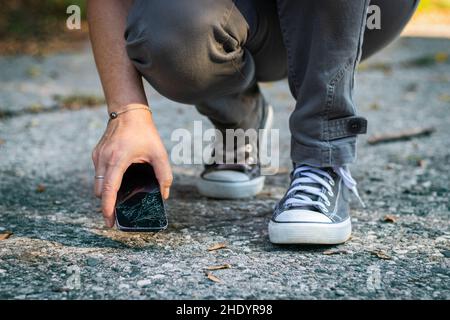 Woman lifting up cracked smartphone from the ground. Damaged mobile phone with cracked touch screen. Stock Photo
