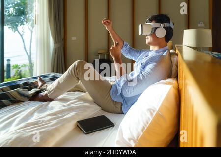 Young Asian man lying on bed and using virtual reality glasses for business meeting during his travel summer vacation holiday. Stock Photo