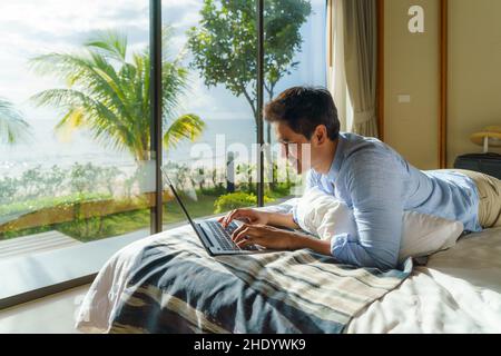 Young Asian businessman lying in bed and working with laptop in room at resort near sea during a summer vacation holiday travel. Stock Photo