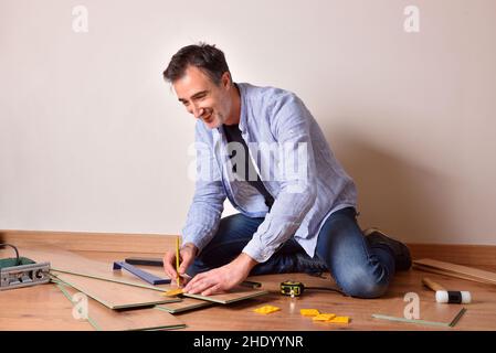 Smiling man sitting on the floor measuring laminate parquet in his own home. Horizontal composition. Stock Photo