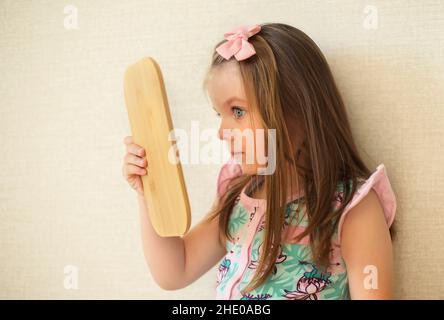 Young curious little girl holding mirror looking at her reflection while playing and spending leisure time at home, wearing makeup and doing it just l Stock Photo