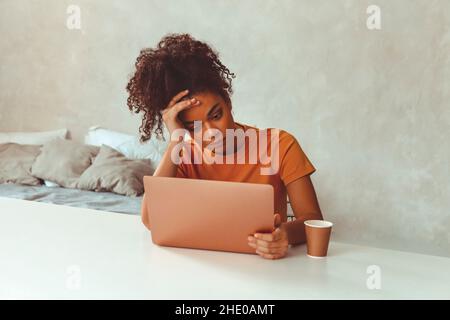 Tired flustrated African descent young girl sitting at desk in front of laptop while irritably looking at computer screen with hands holding her head Stock Photo