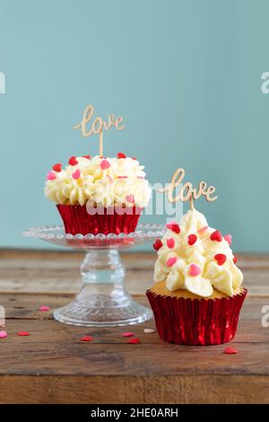 Valentine's Day. Two cupcakes with red topping decoration and wooden sign with the word LOVE on a wooden table with blue background. Vertical photo. Stock Photo