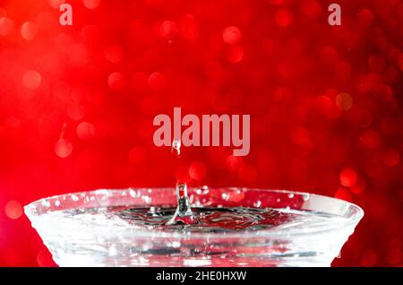 Drop of water falling on filled glass making an isolated splash with a red bokeh background Stock Photo