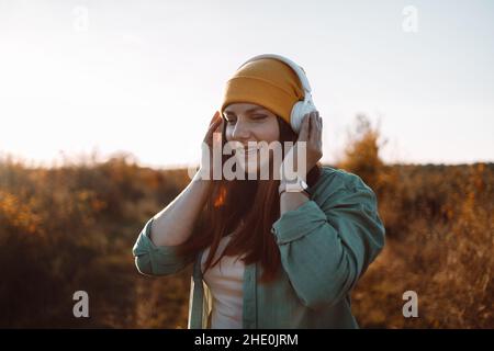 Stylish 30s caucasian female in headphones listening music, enjoys music in the meadow Stock Photo