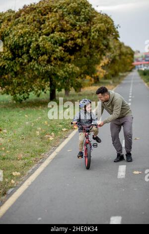 Father teaches his son to ride a bicycle on the bike path in the park. The father is holding a bicycle and the son is sitting on it. Stock Photo