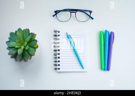 High angle of a white desk with colorful writing instruments on it and a potted green plant, glasses and an empty notebook Stock Photo
