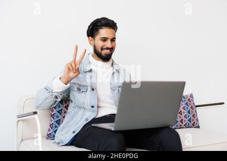 Young happy smiling guy in checkered shirt sitting on sofa at home and using laptop for video conversation Stock Photo