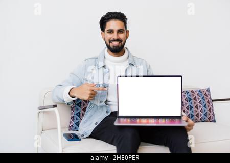 Young indian man lying comfortably on the sofa while using the laptop with blank screen. Stock Photo
