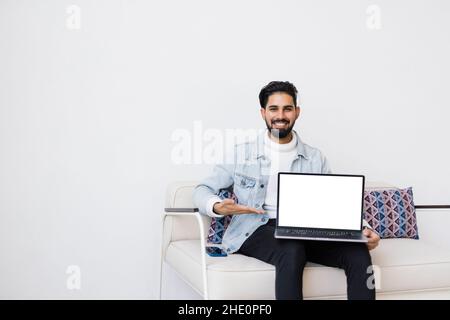Young indian man lying comfortably on the sofa while using the laptop with blank screen. Stock Photo