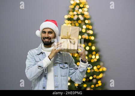 Curious Santa man sitting in front of Christmas tree background. Smiling handsome guy surprised of the present after the opening in the gift box Stock Photo