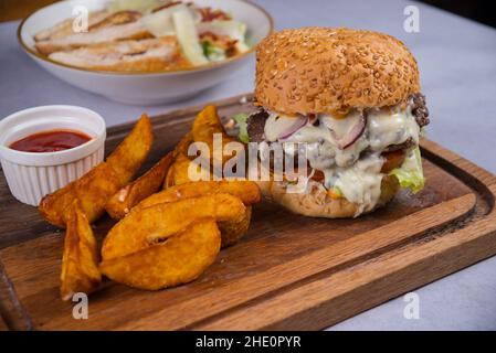 Hamburger with cutlet, cheese and white sauce, potatoes, and tomato sauce on a wooden cutting board Stock Photo