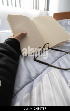 hand of a person holding an open notebook, detail in its blank sheets, next to some glasses, lifestyle and intellectual pastimes, reading Stock Photo