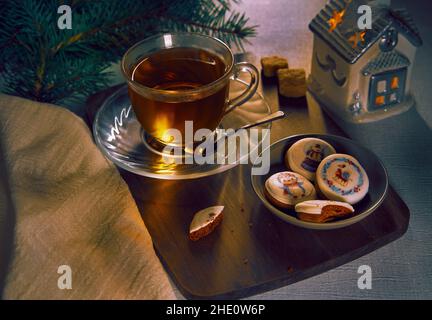 Tea in a transparent cup and gingerbread cookies on a ceramic plate with tree plank with branches of a Christmas tree and toy house in the background Stock Photo