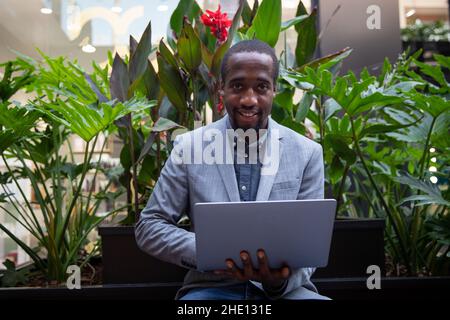 African businessman sitting on a bench works with his laptop. Successful person with confidant look smiles. Businessman works outdoors Stock Photo